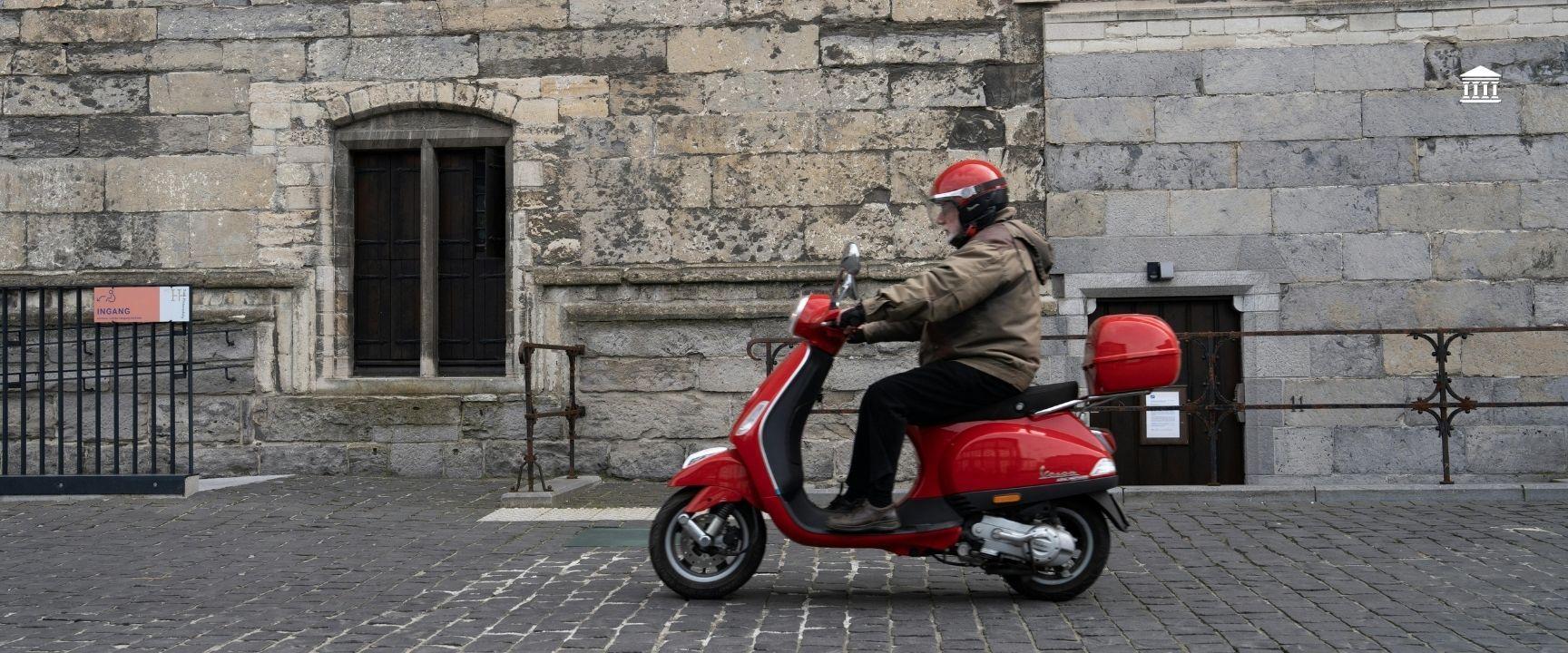 A man on a red scooter in a cobbled street in europe.